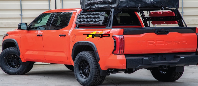 K&ouml;nnte beinhalten: Ein leuchtend orangefarbener Pick-up mit einem schwarzen Dachgep&auml;cktr&auml;ger und einem schwarzen "TRD PRO" Aufkleber auf der Heckklappe. Der Wagen hat schwarze R&auml;der und Reifen.