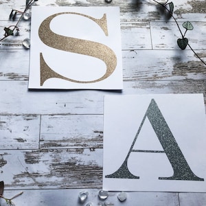K&ouml;nnte beinhalten: Zwei wei&szlig;e Karten mit goldenen und silbernen Glitzerbuchstaben, die Buchstaben sind S und A.
