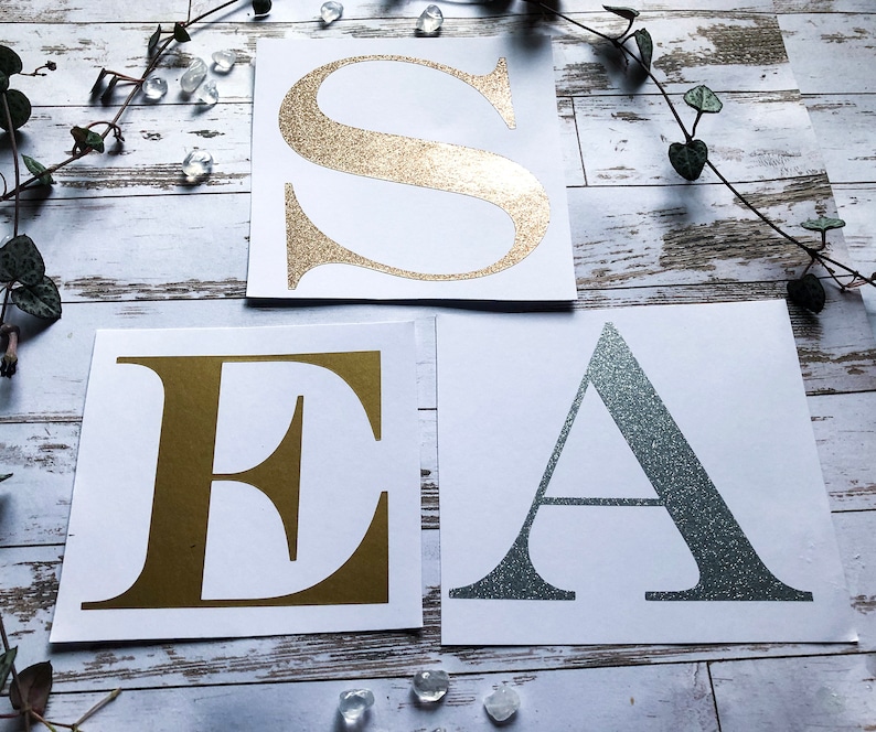 K&ouml;nnte beinhalten: Drei wei&szlig;e Karten mit den Buchstaben "S", "E" und "A" in Gold-, Gold- und Silberglitter. Die Buchstaben befinden sich auf wei&szlig;em Hintergrund.