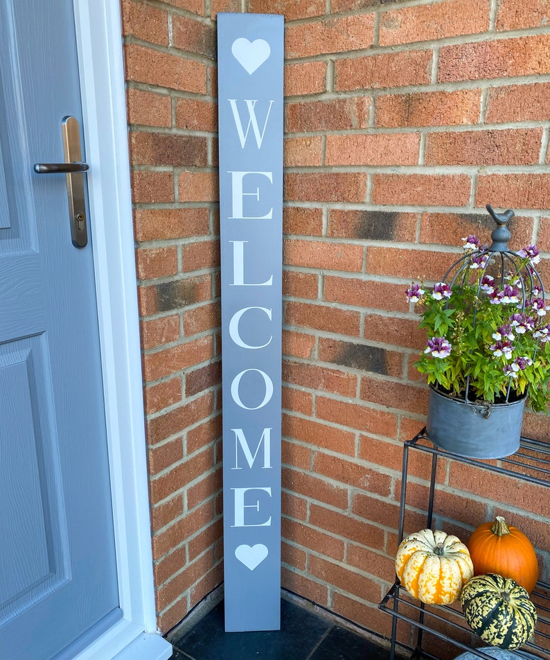 K&ouml;nnte beinhalten: Ein graues Holzschild mit der Aufschrift "Willkommen" in wei&szlig;en Buchstaben und Herzen. Das Schild lehnt an einer Backsteinmauer.