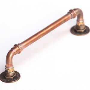 May include: A copper pipe door handle with brass fittings. The handle is curved at both ends and has a brushed copper finish. The brass fittings have a polished finish and are marked with the text "MADE IN UK".