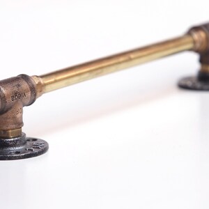 May include: A brass pipe handle with black iron flanges. The handle is about 12 inches long and has a hexagonal nut on one end. The flanges have a decorative pattern.