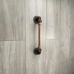 May include: A copper pipe door handle with black metal flanges. The handle is mounted on a light gray wood wall.