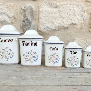Peut inclure: Ensemble de cinq pots de cuisine en céramique blanche avec couvercles. Chaque pot a un bord bleu foncé et est étiqueté en script marron avec des mots français : "Sucre", "Farine", "Café", "Thé", et "Épices". Des motifs floraux roses et bleus décorent le devant.