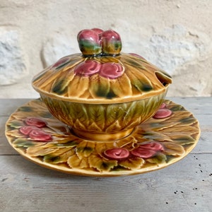 Könnte beinhalten: Ein Keramik-Servierset mit Deckel und passendem Teller. Das Set zeigt ein detailliertes Design aus Rosen und Blättern in Braun-, Grün- und Rottönen. Der Deckel ist mit einem Paar rosenförmiger Griffe versehen.