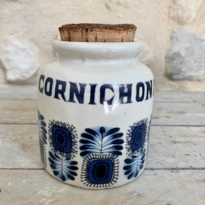 Peut inclure: Un pot en céramique blanc avec le mot "CORNICHON" en lettres bleues. Le pot présente un motif floral bleu répétitif et est surmonté d'un bouchon en liège. Le pot est probablement destiné à conserver des cornichons ou autres conserves.