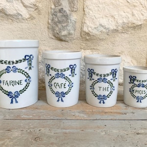 Könnte beinhalten: Ein Set aus vier weißen Keramikbehältern in verschiedenen Größen, jeweils mit einem blau-grünen Lorbeerkranz-Motiv und Schleifen verziert. Die Behälter sind mit dem schwarzen Text FARINE, CAFÉ, THÉ und EPICES in einem Oval beschriftet.