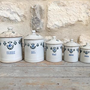 Peut inclure: Un ensemble de cinq pots en céramique blanche avec couvercles, disposés par taille décroissante. Chaque pot présente un motif floral bleu sur le haut et les mots FARINE, SEL, THÉ, POIVRE et ÉPICES inscrits en bleu sur la face avant.
