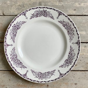 Peut inclure: Assiette en céramique blanche avec un motif floral violet sur le bord. L'assiette a un large bord avec une bordure à carreaux noirs et blancs. Le motif floral comprend des fleurs et des feuilles stylisées.