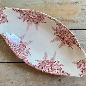May include: An oval serving dish with a white base and a burgundy floral pattern. The dish has a scalloped edge and is decorated with flowers and ornate scrollwork. The dish is placed on a weathered wooden surface.