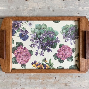 May include: Rectangular wooden serving tray with a floral design. The tray has a light brown wooden frame and two semi-circular handles. The floral design features purple, pink, and yellow flowers on a cream background.