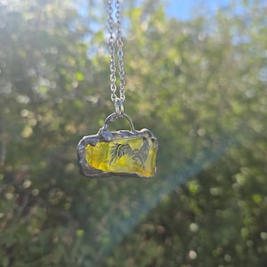May include: A rectangular pendant necklace featuring a translucent yellow glass piece with a black bird illustration inside. The glass is encased in a textured silver-toned metal frame with a small loop at the top, suspended from a silver-toned chain.
