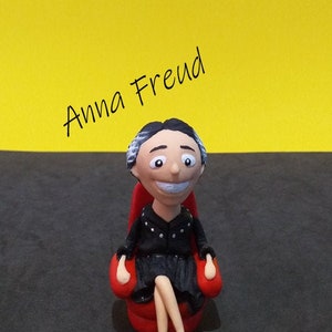 Könnte beinhalten: Eine handbemalte Figur von Anna Freud, einer Psychoanalytikerin, die in einem roten Sessel sitzt. Die Figur ist aus Polymer Clay gefertigt und hat einen skurrilen, cartoonartigen Stil. Der Text "Anna Freud" ist in Schwarz über der Figur geschrieben.