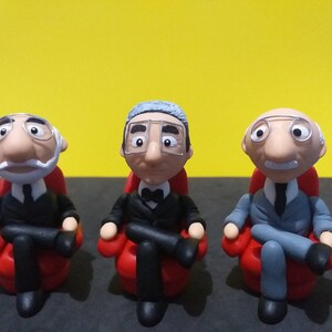 May include: Three small figurines of men wearing suits and glasses, sitting in red chairs. The men have grey hair and are smiling.