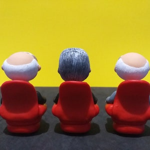 May include: Three small figurines of people sitting in red chairs, facing a yellow wall. The figurines are bald, with grey hair, and have white faces.