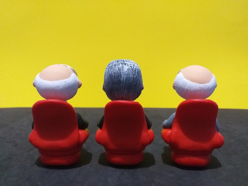 May include: Three small figurines of people sitting in red chairs, facing a yellow wall. The figurines are bald, with grey hair, and have white faces.