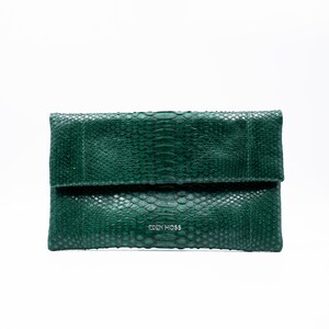 Könnte beinhalten: Dunkelgrüne Clutch mit Schlangenmuster. Die Clutch hat eine Klappe zum Zusammenfalten und den Markennamen "EDEN MOSS". Das Material ist strukturiert, vor weißem Hintergrund.