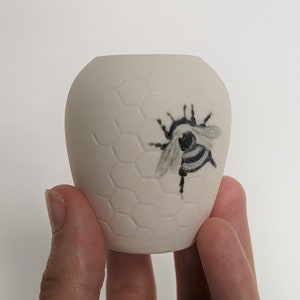 Puede incluir: Un pequeño jarrón de cerámica blanca con un patrón de panal y una ilustración de abeja en blanco y negro.