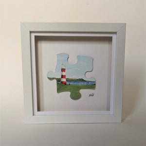Pu&ograve; includere: Una cornice bianca con un passepartout bianco che circonda un pezzo di puzzle di faro a strisce rosse e bianche. Il faro si trova su un'area erbosa verde con un'estensione d'acqua blu sullo sfondo. Il pezzo di puzzle &egrave; firmato "Molloy".