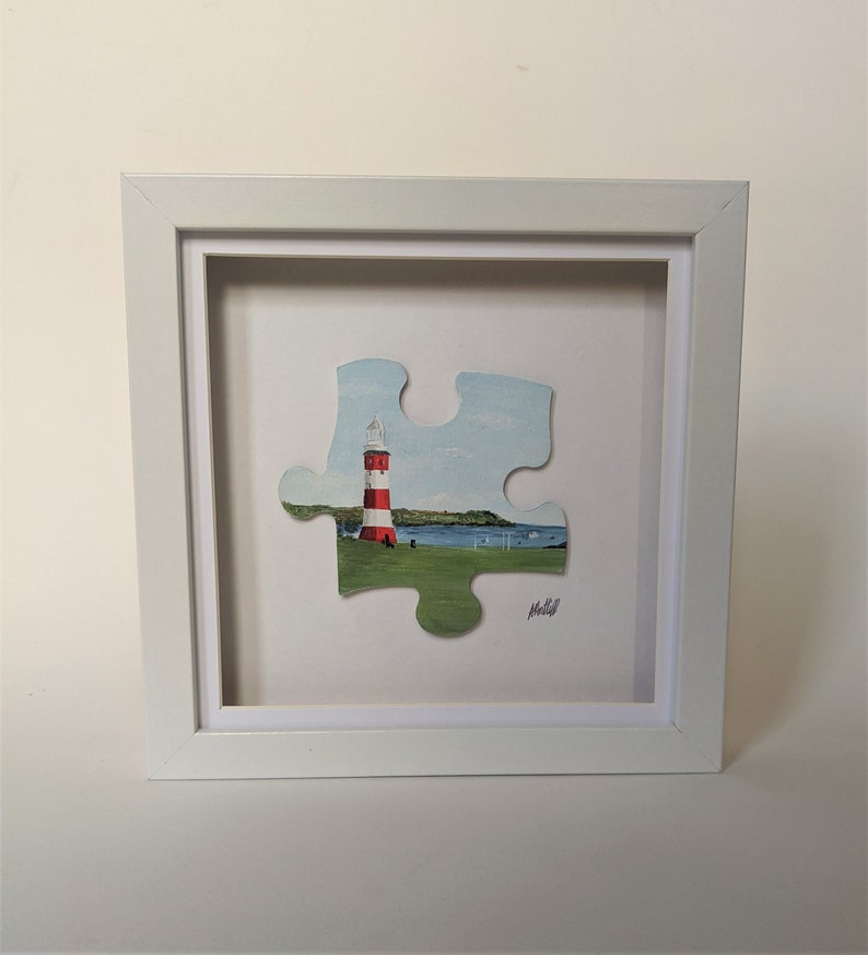 Pu&ograve; includere: Una cornice bianca con un passepartout bianco che circonda un pezzo di puzzle di faro a strisce rosse e bianche. Il faro si trova su un'area erbosa verde con un'estensione d'acqua blu sullo sfondo. Il pezzo di puzzle &egrave; firmato "Molloy".