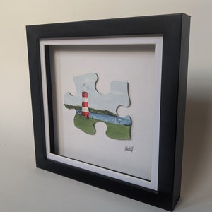 Pu&ograve; includere: Un dipinto incorniciato di un faro rosso e bianco su un pezzo di puzzle verde e blu. Il pezzo &egrave; rialzato rispetto allo sfondo bianco e il dipinto &egrave; firmato "Alistif".