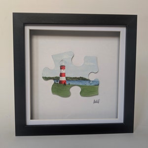 Pu&ograve; includere: Un dipinto incorniciato di un faro rosso e bianco su una collina verde erbosa con un'acqua in fondo. Il dipinto ha la forma di un pezzo di puzzle.