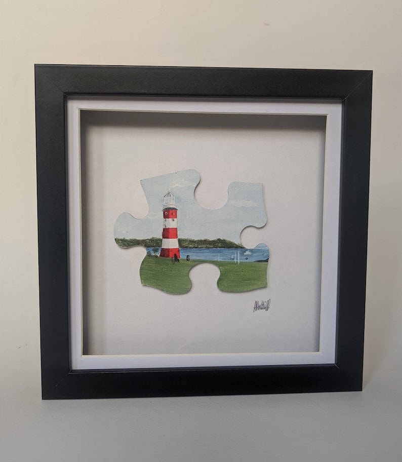 Pu&ograve; includere: Un dipinto incorniciato di un faro rosso e bianco su una collina verde erbosa con un'acqua in fondo. Il dipinto ha la forma di un pezzo di puzzle.