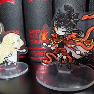 May include: Two anime-style character figurines on clear acrylic stands. The figurine on the left is a blonde woman with a white dress and a golden staff. The figurine on the right is a red-haired woman with a black and red outfit, a red snake, and black wings.