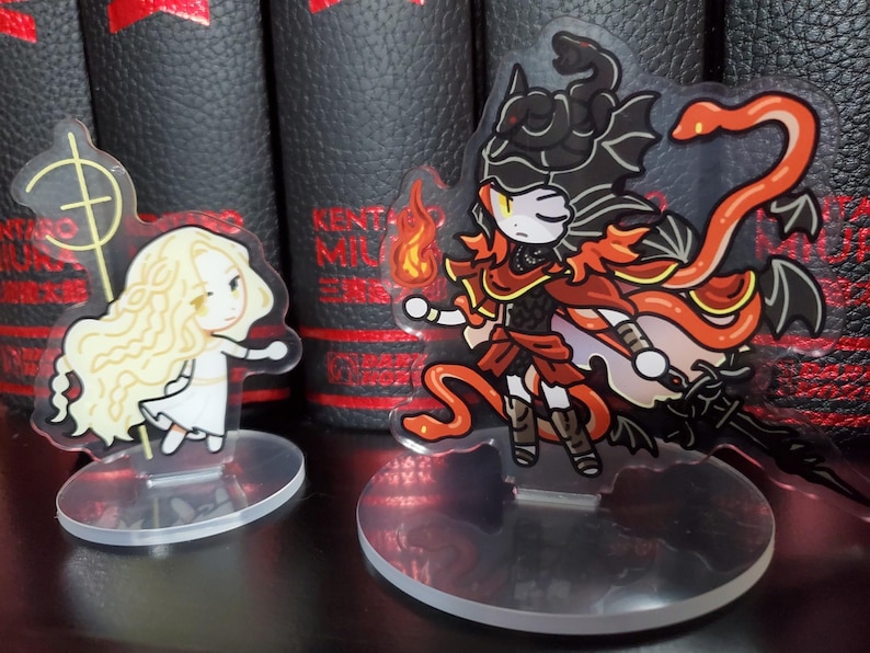 May include: Two anime-style character figurines on clear acrylic stands. The figurine on the left is a blonde woman with a white dress and a golden staff. The figurine on the right is a red-haired woman with a black and red outfit, a red snake, and black wings.