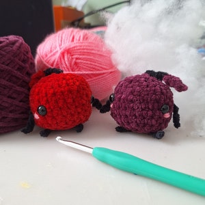 May include: Two small crocheted plush toys in the shape of bugs. One is red with black legs and the other is purple with black legs. Both have black eyes and pink cheeks.