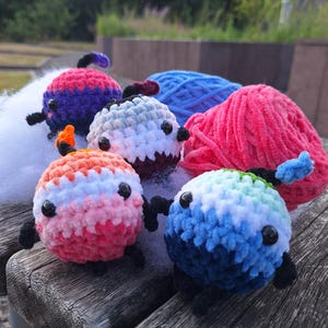 May include: A collection of colorful, crocheted plush toys, each with a round body and small black features. The toys are in shades of pink, orange, blue, and purple, with black appendages. A ball of pink and blue yarn is in the background.