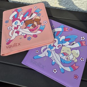 Puede incluir: Dos carpetas cuadradas para tarjetas coleccionables, una rosa y otra morada, cada una con el logotipo de Vaultx y un gráfico del Pokémon Sylveon interactuando con Eevee, rodeados de pequeñas flores blancas y azules y detalles de corazones.