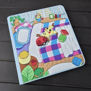 May include: A hand-painted, light blue binder depicting a bedroom scene. The artwork includes a bed with a checkered blanket, a Mimikyu character, a Pokeball, books, and a window with curtains. The binder has a stitched border.
