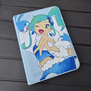 May include: A light blue rectangular binder featuring a printed anime-style character with teal hair, a blue outfit with white fur trim, and a small blue creature. The character is winking and reaching forward. The word vault is visible at the bottom.