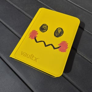 May include: Yellow card holder with a cartoon face design. The card holder has a black zig-zag mouth, black scribbled eyes, and red cheek accents. The text "VaultX" is printed in the lower left corner.