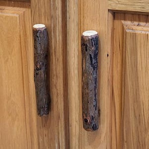 Może przedstawiać: Drewniane drzwi szafki z rustykalnymi uchwytami w stylu gałązek. Drzwi szafki wykonane są z jasnego drewna z widocznym usłojeniem. Uchwyty w kształcie gałązek są ciemnobrązowe i cylindryczne, dodając naturalny element.