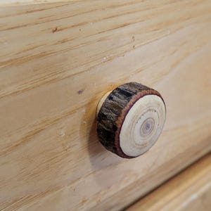 Könnte beinhalten: Ein hölzerner Schrankknauf aus einem Querschnitt eines Astes. Der Knauf hat ein natürliches Holzmaserungsmuster mit hellbrauner Farbe und einem dunkelbraunen Rindenrand. Er ist an einem hellfarbigen Holzschrank befestigt.