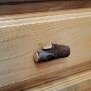 May include: A close-up of a wooden drawer with a natural twig-shaped handle. The drawer is made of light-colored wood with visible grain, and the handle is a dark brown color. The handle is approximately 3 inches long.