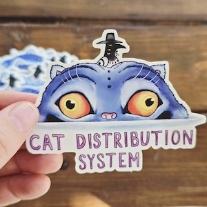 Peut inclure: Autocollant représentant un chat bleu aux grands yeux orange et un petit oiseau portant un chapeau. Le texte "CAT DISTRIBUTION SYSTEM" est en dessous du chat. L'autocollant est tenu sur un fond en bois.