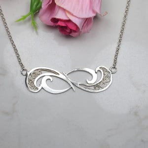 Könnte beinhalten: Eine silberfarbene Halskette mit einem Unendlichkeitszeichen-Anhänger. Der Anhänger weist aufwendige Filigranarbeiten auf und hängt an einer zarten Kette. Die Halskette wird auf einer Marmoroberfläche mit einer rosa Blume im Hintergrund präsentiert.