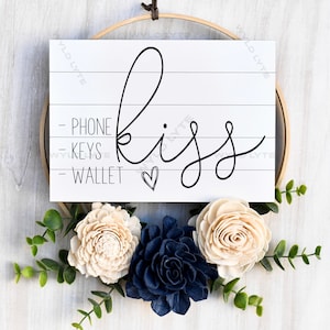 May include: White wooden sign with the word "kiss" written in black cursive script. The sign has three lines of text below the word "kiss" that read "phone", "keys", and "wallet". The sign is surrounded by white wood and greenery.
