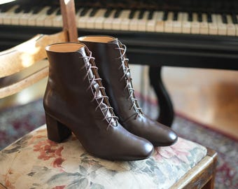 May include: A pair of brown leather ankle boots with a lace-up design and block heels. The boots are placed on a floral patterned chair, with a piano in the background.