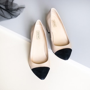 May include: A pair of beige heeled shoes with black toe caps. The shoes have a pointed toe and a low block heel. The brand name "Anna Marziali" is printed on the inside of each shoe.