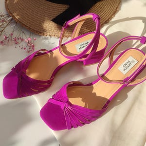 Może przedstawiać: Para fuksjowych sandał&oacute;w z paskami na kostkę i niskim obcasem. Sandały mają kwadratowy nosek i wiązany wz&oacute;r na cholewce. Marka "ANNA MARATTI" jest widoczna na wkładce. Słomkowy kapelusz i r&oacute;żowe kwiaty są w tle.