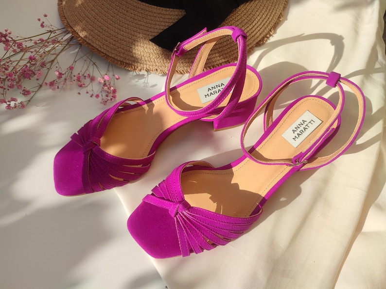 Może przedstawiać: Para fuksjowych sandał&oacute;w z paskami na kostkę i niskim obcasem. Sandały mają kwadratowy nosek i wiązany wz&oacute;r na cholewce. Marka "ANNA MARATTI" jest widoczna na wkładce. Słomkowy kapelusz i r&oacute;żowe kwiaty są w tle.