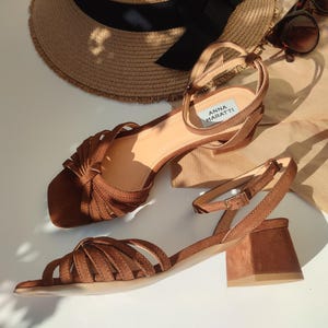 Puede incluir: Sandalias de ante marr&oacute;n con correas en el tobillo y tacones cuadrados. Las sandalias tienen m&uacute;ltiples correas en los dedos y una peque&ntilde;a etiqueta que dice "ANNA MARATTI". Un sombrero de paja con una cinta negra y gafas de sol tambi&eacute;n est&aacute;n en la imagen.