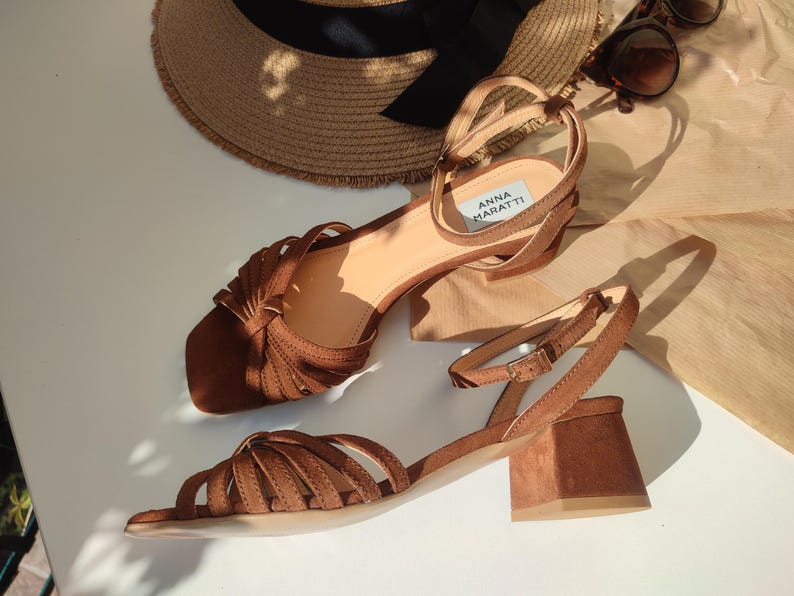 Puede incluir: Sandalias de ante marr&oacute;n con correas en el tobillo y tacones cuadrados. Las sandalias tienen m&uacute;ltiples correas en los dedos y una peque&ntilde;a etiqueta que dice "ANNA MARATTI". Un sombrero de paja con una cinta negra y gafas de sol tambi&eacute;n est&aacute;n en la imagen.