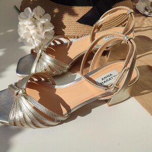 May include: A pair of gold-colored sandals with a square toe and ankle strap. The sandals have multiple straps across the top and a small heel. The brand name "ANNA MARATTI" is visible on the side of one sandal. The sandals are approximately 2 inches in heel height.