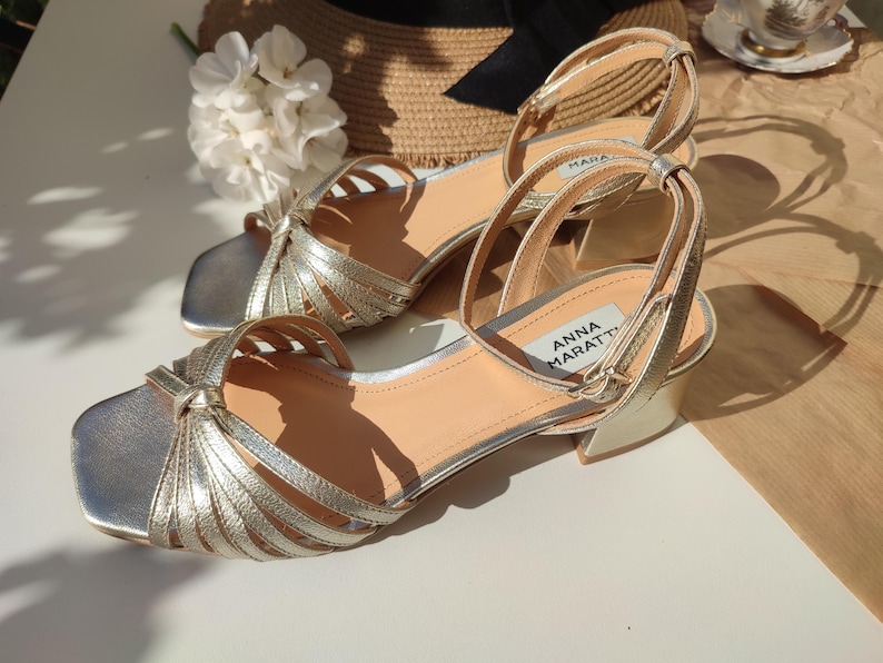 May include: A pair of gold-colored sandals with a square toe and ankle strap. The sandals have multiple straps across the top and a small heel. The brand name "ANNA MARATTI" is visible on the side of one sandal. The sandals are approximately 2 inches in heel height.