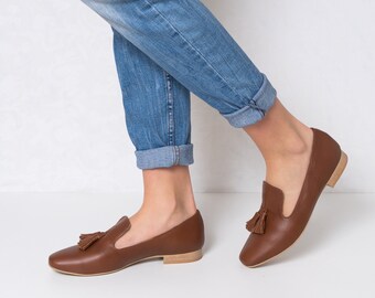 Puede incluir: Mocasines de cuero marrón con borla. Los zapatos tienen un diseño clásico con tacón bajo y punta redonda. Los mocasines se combinan con unos vaqueros azules. Los zapatos parecen estar hechos de cuero de alta calidad.